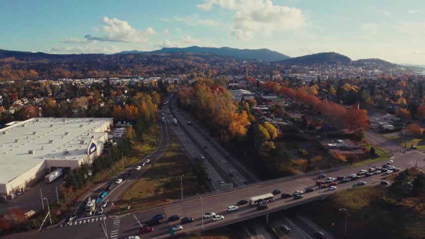 An aerial view shows a busy highway interchange with an overpass and flowing traffic through a suburban landscape with vibrant autumn foliage and distant hills
