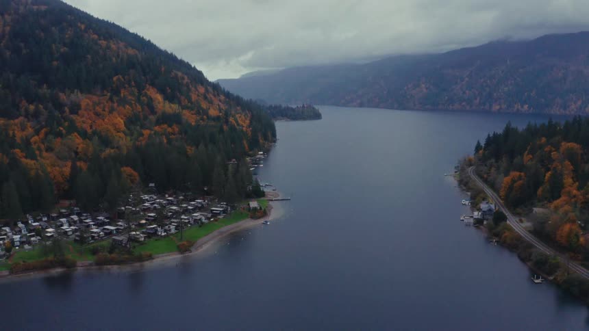 An expansive aerial view of a mountain lake reveals a quiet shoreline community amidst colorful autumn forests on a cloudy day
