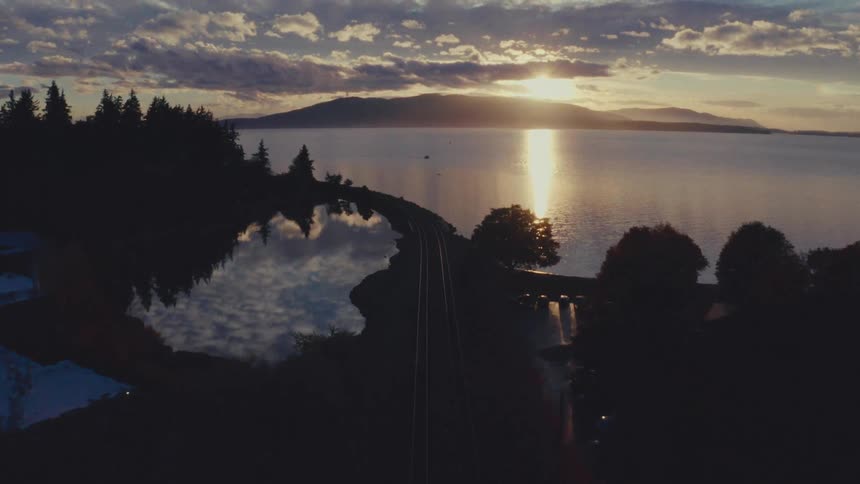 A serene aerial shot follows railroad tracks along a silhouetted coastline toward a brilliant sunrise reflecting on the water