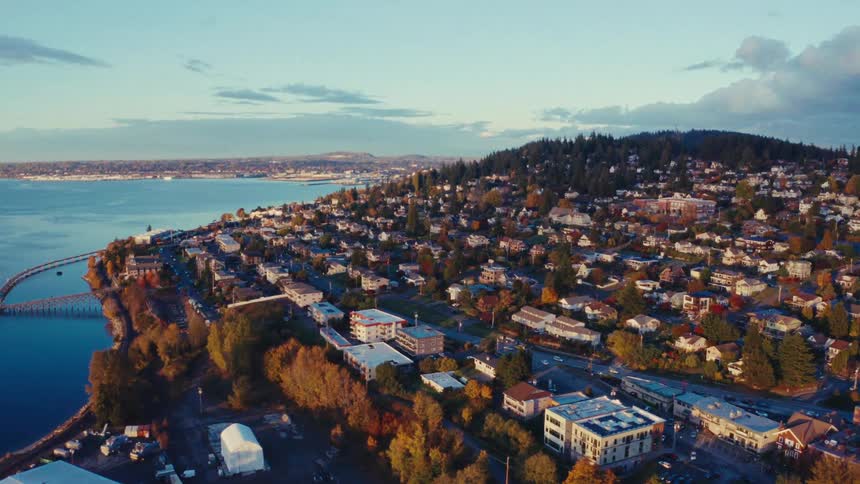 An aerial view slowly panning right over a picturesque coastal town nestled on a forested hill during a vibrant autumn sunset
