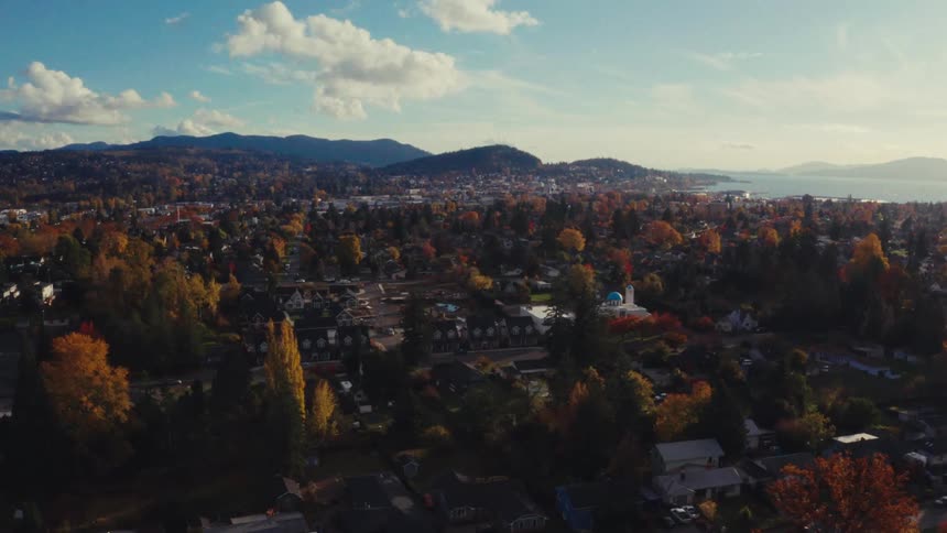 An aerial view of a picturesque coastal town during autumn, with colorful trees, distant mountains, and a bright blue sky