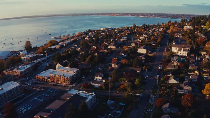 A slow aerial ascent reveals a peaceful coastal town with colorful autumn trees waking up to a golden sunrise