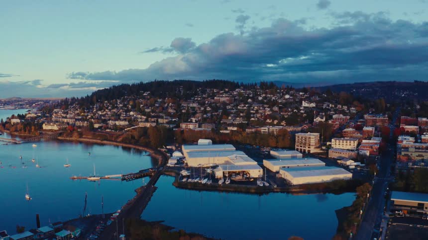 An aerial view flies over a waterfront industrial area towards a beautiful coastal town on a hill at sunrise