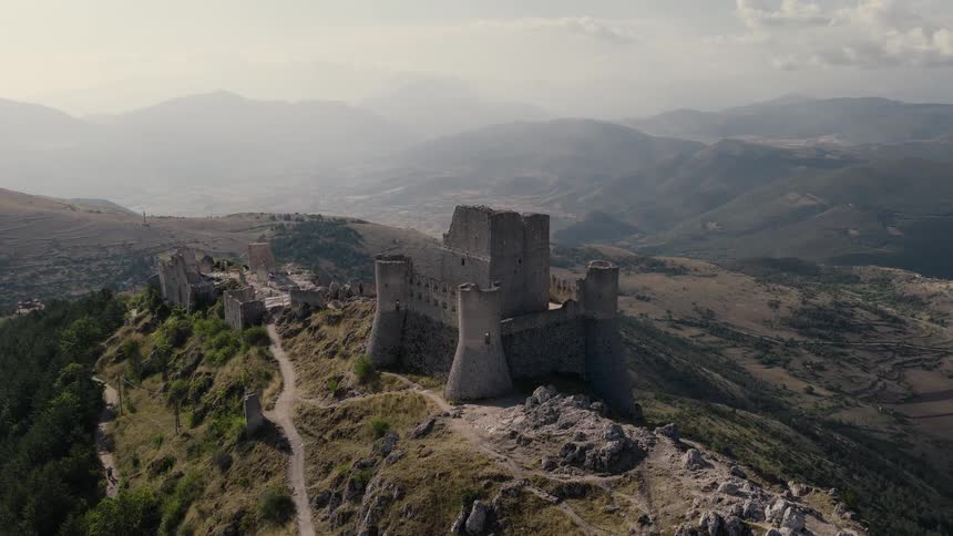 An epic aerial shot orbits the ancient, ruined stone castle of Rocca Calascio perched atop a dramatic mountain in Abruzzo, Italy