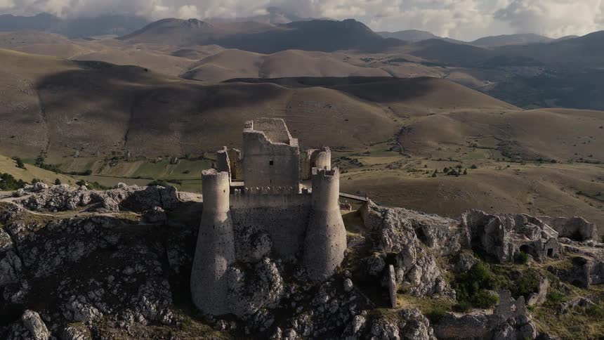 A slow, cinematic aerial orbit reveals the ancient Rocca Calascio castle ruins standing majestically on a rocky peak amidst the rolling hills of Abruzzo, Italy