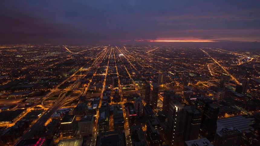 A high-angle time-lapse of a sprawling city grid as a vibrant sunset fades and the urban landscape illuminates with millions of lights