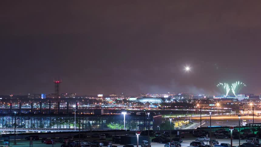 Time-lapse of nighttime airport traffic with colorful fireworks exploding in the distance over the city