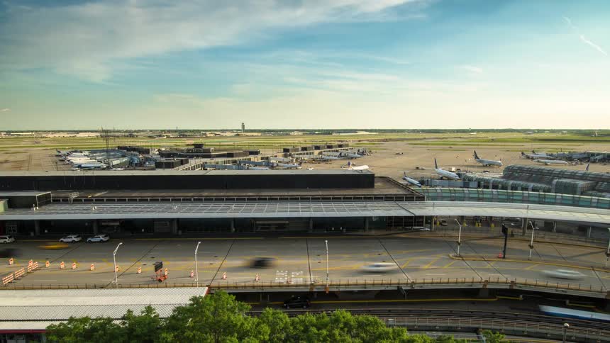 Wide-angle time-lapse of a bustling airport terminal and runways, with cars and trains zipping by in the foreground during the day