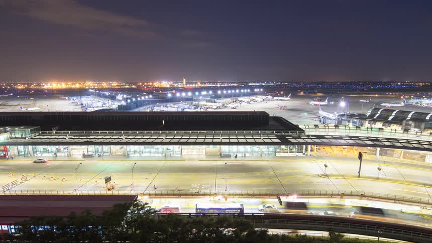 A bustling international airport comes to life in a wide timelapse at night, showing busy traffic and airplanes on the brightly lit tarmac