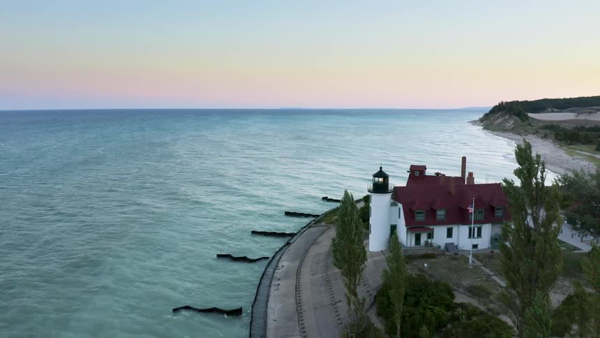 A scenic aerial view slowly approaches the Point Betsie Lighthouse on the shores of Lake Michigan at sunset