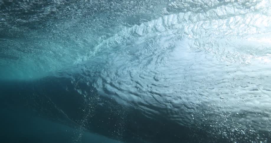 A slow-motion underwater view captures a powerful ocean wave swelling and passing overhead, with sunlight illuminating the turbulent water