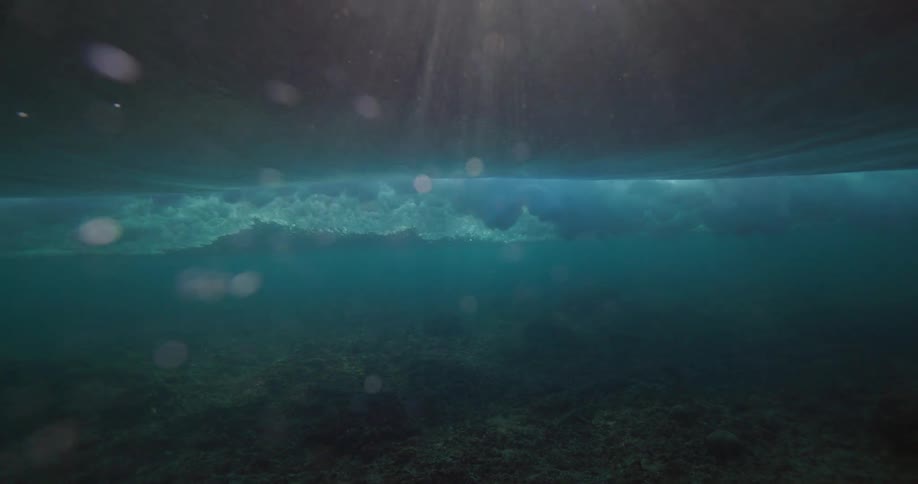 A stunning slow-motion underwater view shows a turquoise ocean wave breaking, sending bubbles through the water as sunlight streams from above