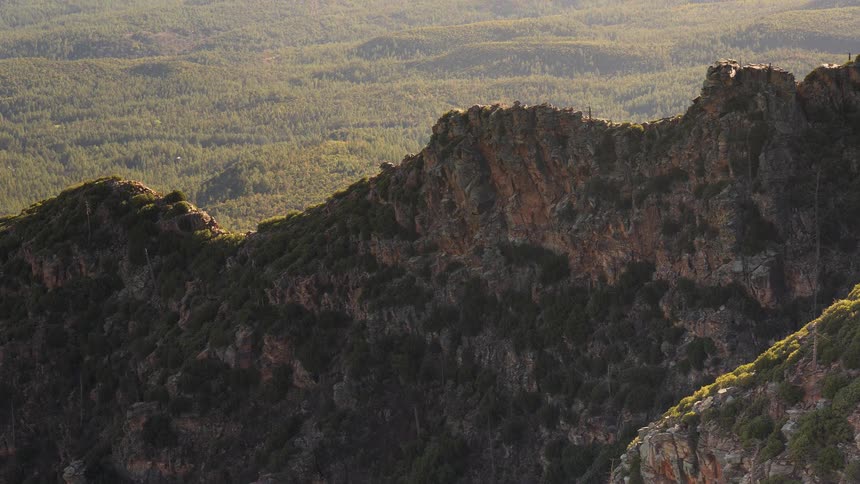 A slow zoom-out reveals a vast, rolling forest landscape beyond a rugged, sunlit mountain ridge