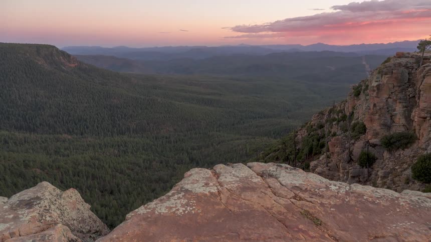 A time-lapse of the sun setting over a vast, forested mountain valley, viewed from the edge of a rocky cliff as the sky turns from gold to pink and purple