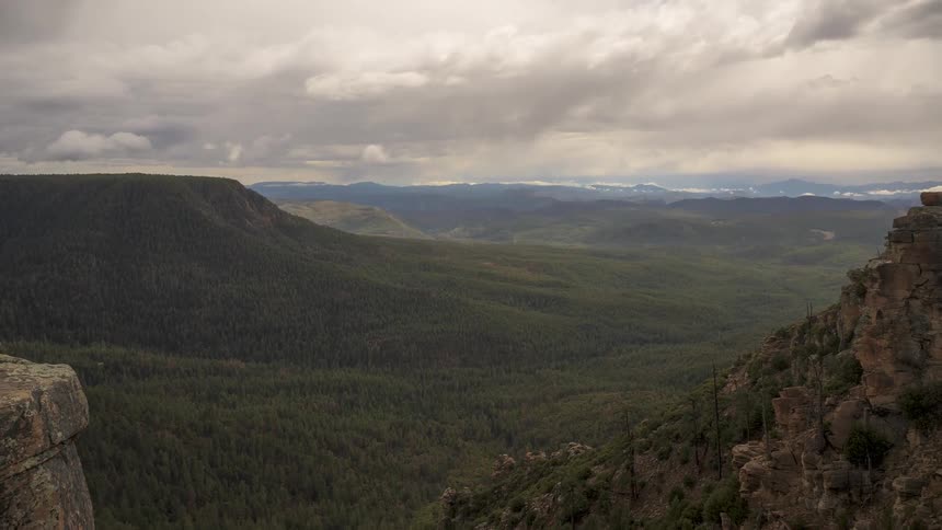 Slowly pulling back from a dramatic, cloudy sky to reveal an expansive forested canyon landscape from a rocky overlook