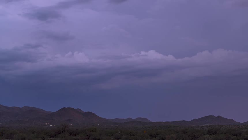 A dramatic time-lapse shows a powerful lightning storm flashing within purple clouds over a dark mountain range at dusk