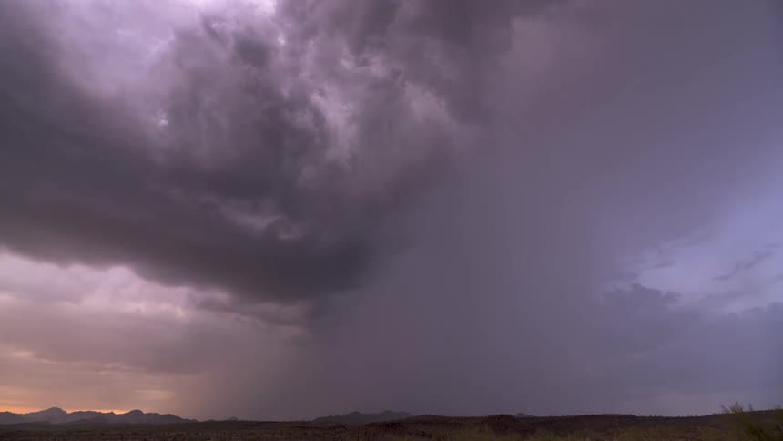 A powerful thunderstorm with internal lightning rolls over a vast desert landscape at dusk as rain begins to fall