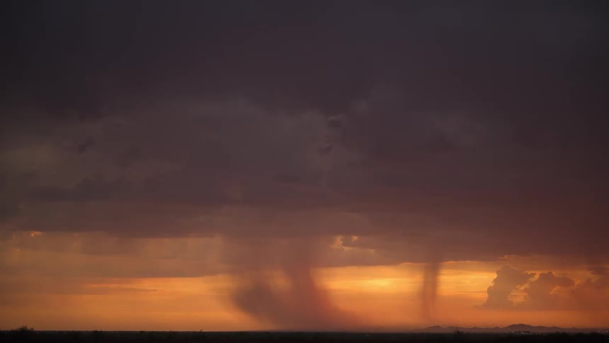 Rain shafts pour from dark, dramatic storm clouds illuminated by a vibrant orange sunset