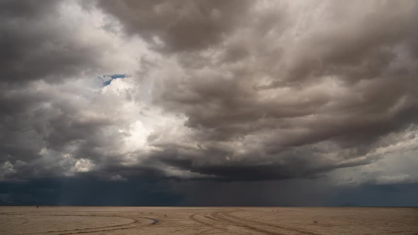 Ominous storm clouds swirl and rain falls in the distance over a dry desert lakebed in this time-lapse