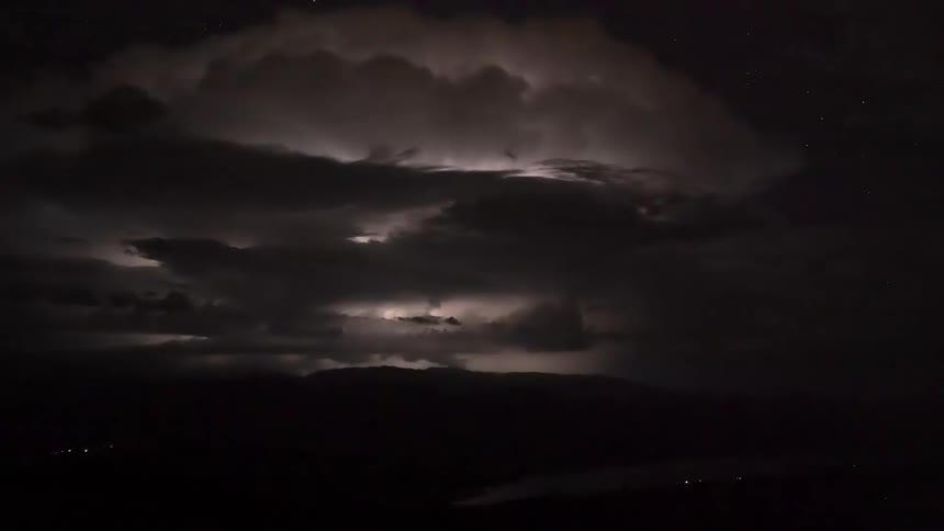 A dramatic time-lapse shows a massive thunderstorm with intense internal lightning flashing over mountains and a lake at night