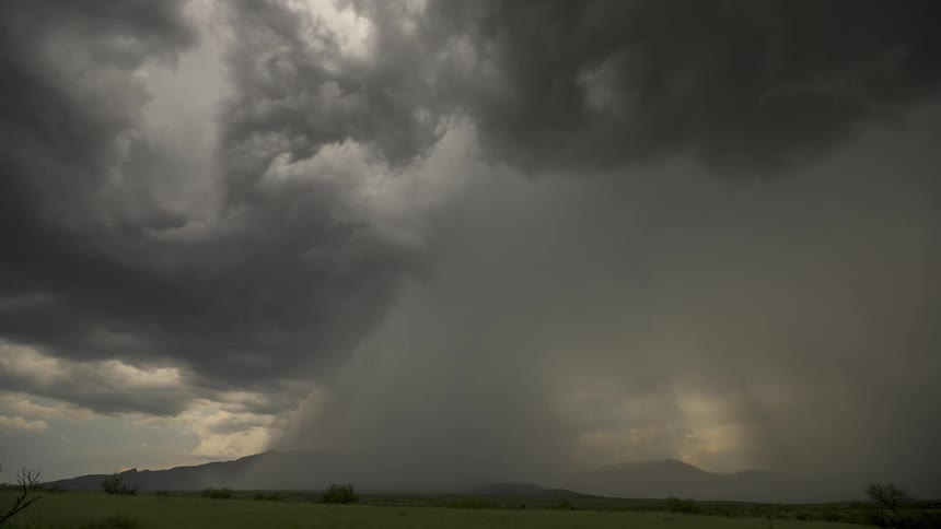 An intense thunderstorm with dark, churning clouds unleashes a heavy downpour over a distant mountain range and green valley