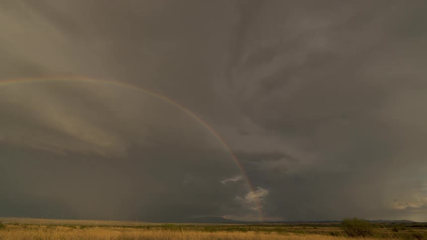 A beautiful time-lapse captures a double rainbow emerging from dark storm clouds, punctuated by a dramatic lightning strike over a golden grassland