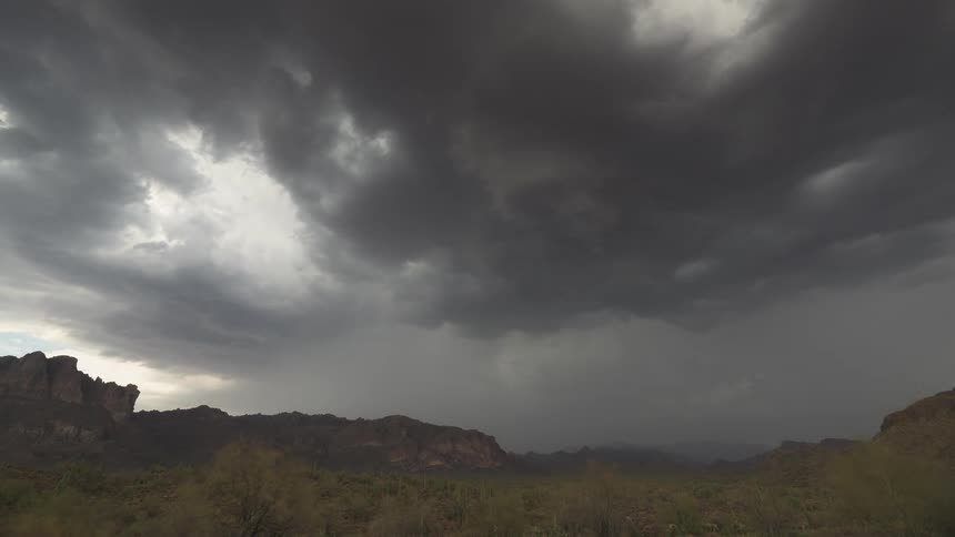 Dark, ominous storm clouds swirl and release rain over a vast desert mountain landscape