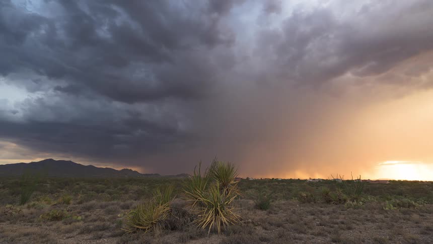 A dramatic desert landscape features dark storm clouds and falling rain illuminated by a beautiful orange sunset
