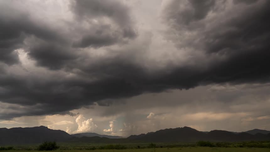 Dark, dramatic storm clouds release a downpour of rain over a mountain range and a vast, green valley