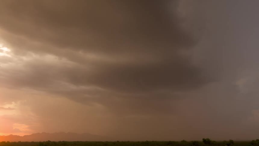 A dramatic time-lapse captures a massive thunderstorm with sheets of rain moving over a desert mountain range during a golden sunset