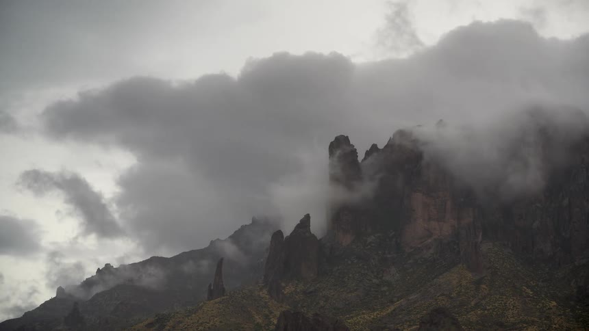 A dramatic timelapse shows moody clouds and fog swirling around the jagged peaks of a rugged mountain range