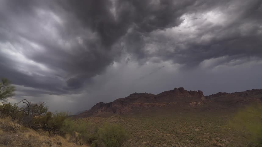 Dramatic dark storm clouds churn and billow over a vast desert mountain landscape as rain falls in the distance