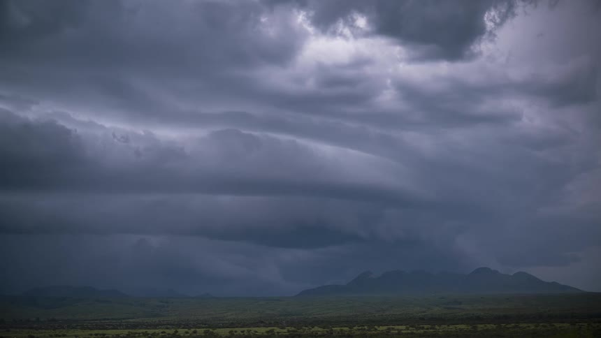 A dramatic timelapse shows dark, ominous storm clouds rolling and swirling over a distant mountain range and a vast green landscape