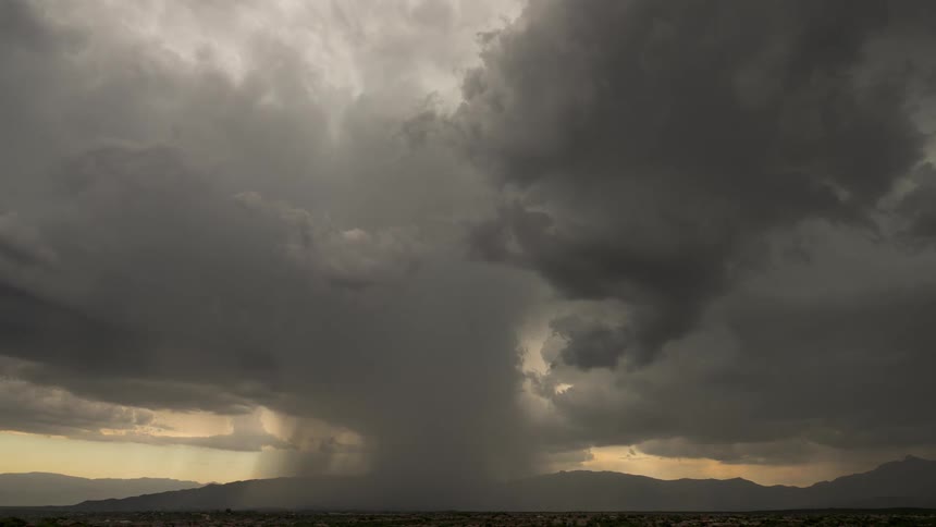 A powerful microburst unleashes a torrent of rain from dark, swirling storm clouds over a distant mountain landscape as light rays emerge
