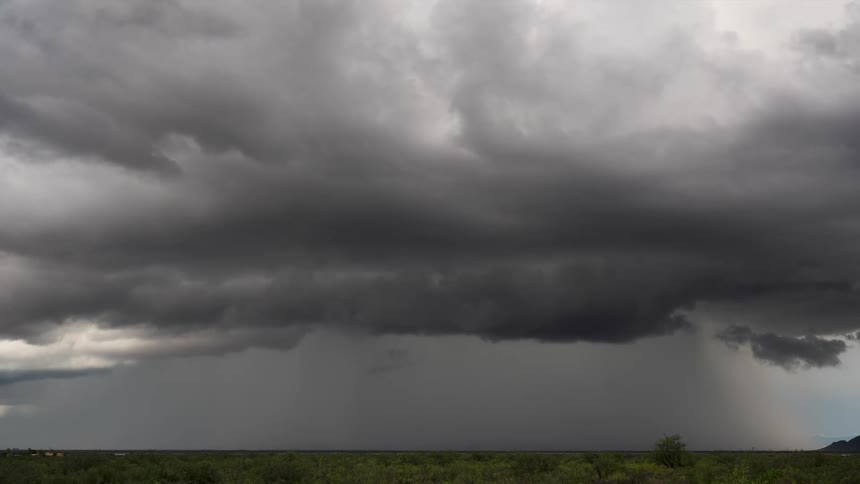 Time-lapse of a massive, dark supercell storm cloud unleashing a powerful downpour over a flat, green plain