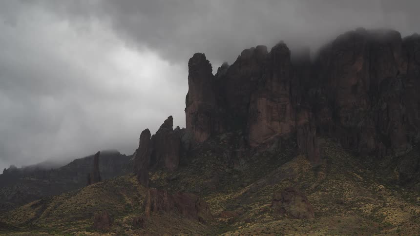 Ominous storm clouds and fog pass over the jagged peaks of a desert mountain range