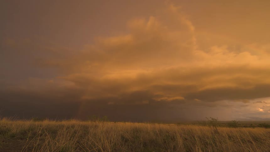 A vibrant rainbow arcs across a dramatic, golden sunset sky over a grassy field before fading away