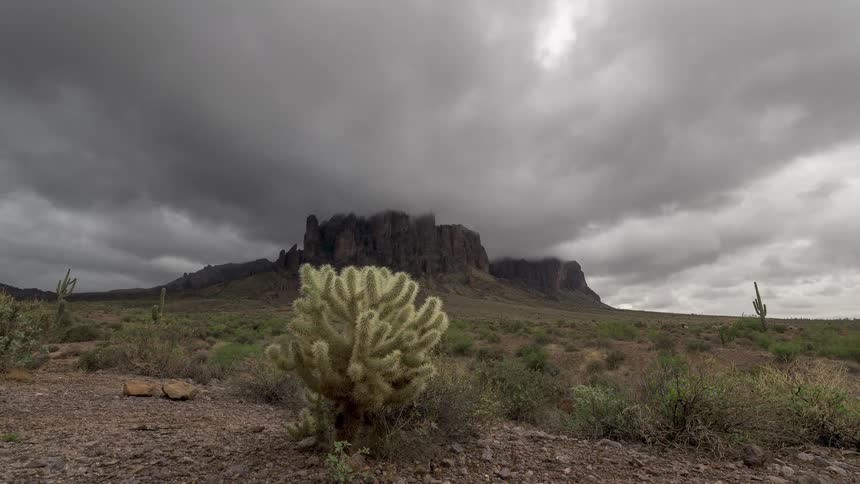 Dramatic time-lapse shows ominous storm clouds rolling over a rugged desert mountain range behind a Cholla cactus