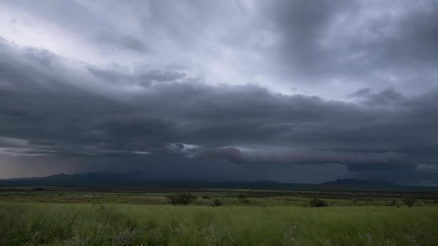 A dramatic timelapse shows a powerful thunderstorm moving over a green, windy field, with the sunset illuminating the dark clouds in vibrant pink and purple hues