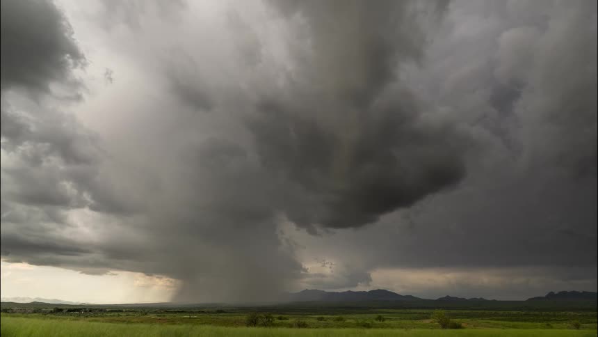 Ominous storm clouds swirl and release rain over a vast green meadow with mountains in the distance