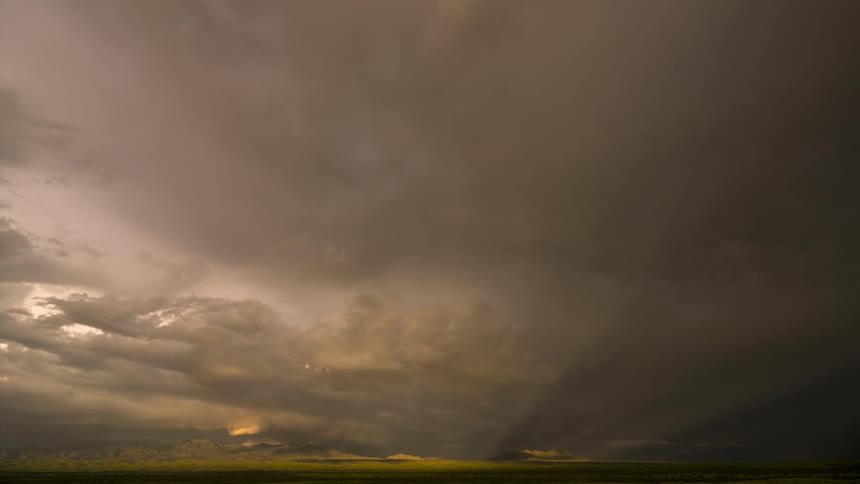 A dramatic time-lapse captures a faint rainbow and golden sunset rays breaking through dark storm clouds over a vast, sunlit plain