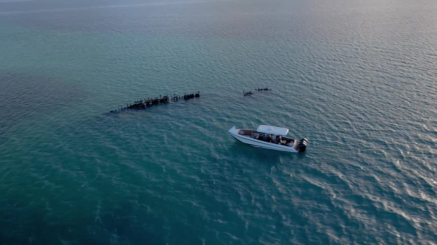 An aerial view shows a tour boat floating near a sunken shipwreck where birds are perched in the clear turquoise ocean