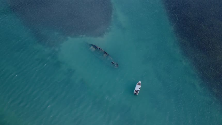 Top-down aerial view of a tourist boat floating next to a large shipwreck submerged in clear turquoise water