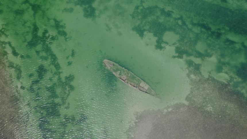 A static top-down aerial view of a shipwreck submerged in shallow, clear green water