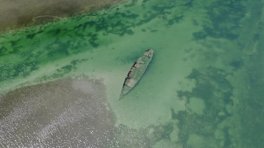 An aerial top-down view reveals a historic shipwreck submerged in the calm, emerald-green sea