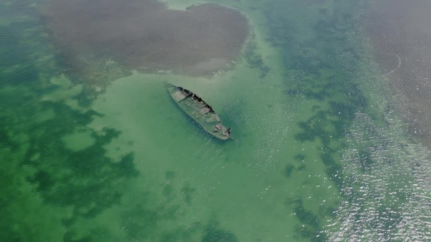 A slow ascending aerial view reveals a sunken shipwreck resting just below the surface in clear, shallow green water
