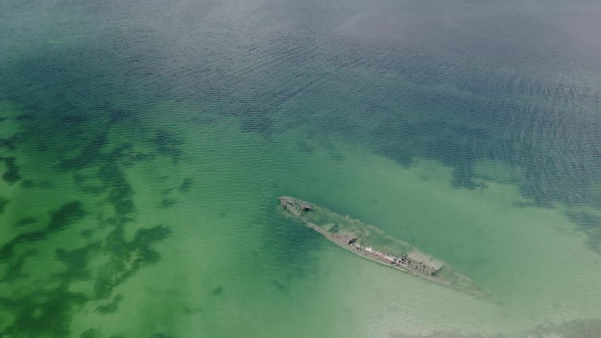 An aerial view slowly tilts down over a shipwreck resting in the shallow, clear green water of the sea