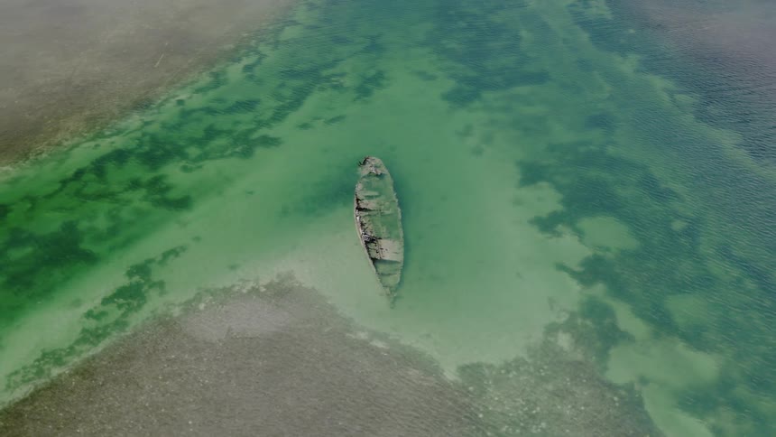 An eerie top-down aerial view slowly zooms out from a sunken shipwreck resting in shallow, clear green water