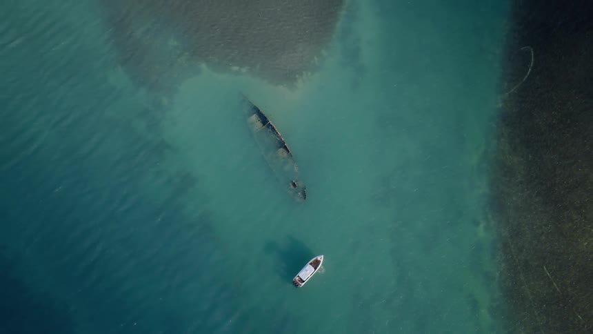 A static aerial top-down shot shows a small boat anchored near a shipwreck submerged in clear, turquoise water