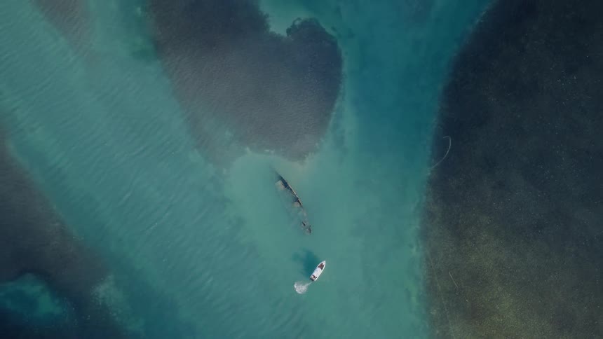 Aerial top-down view of a fisherman casting a net from a small boat next to a shipwreck in shallow turquoise water